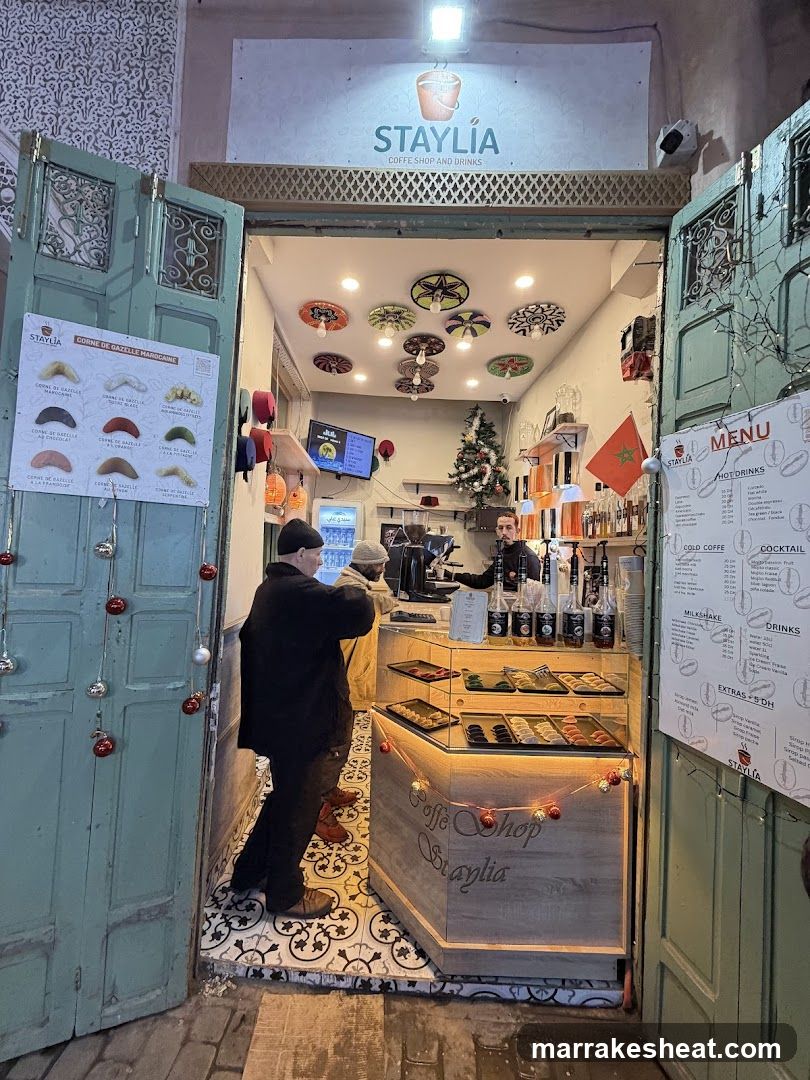 The image shows the entrance of Staylia, a coffee shop and drinks establishment. The storefront features a sign with the name 'STAYLIA' above the doorway. Inside, there is a counter displaying various food items under glass cases. A person wearing a black coat stands near the counter, while another individual appears to be working behind it. Decorative elements include colorful circular wall hangings and string lights around the display case. To the left side of the frame, a menu board lists different types of pastries or snacks with corresponding prices.