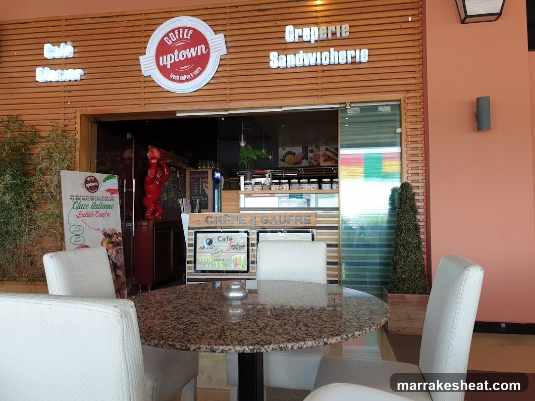 The image shows the storefront of a cafe and sandwich shop with wooden slat walls. Visible signage includes 'Coffee Uptown' in a circular logo, 'Creperie', and 'Sandwicherie'. A glass entrance reveals the interior counter area. Outside, there is a round granite table surrounded by white chairs. To the left, a promotional poster advertises Italian ice cream flavors. Inside, red statues and potted plants are visible near the counter.