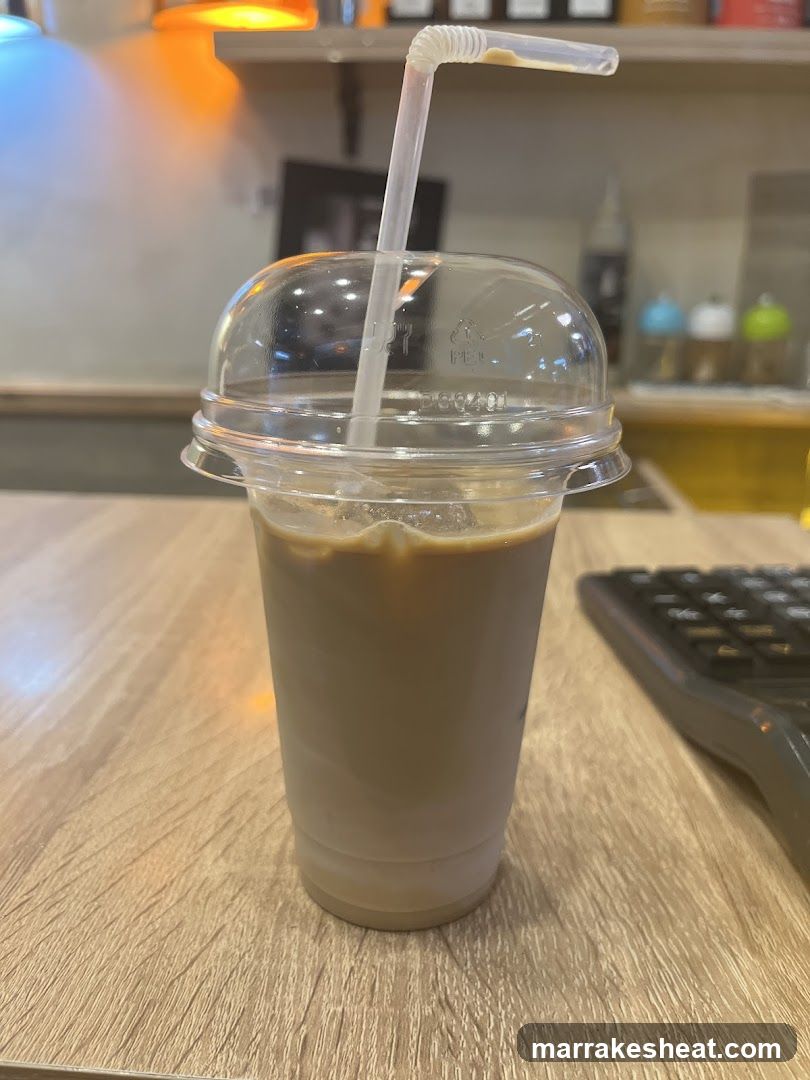 A clear plastic cup with a dome lid and flexible straw containing iced coffee, placed on a wooden table. In the background, there is a blurred interior of a shop or cafe with shelves holding various items and warm lighting.