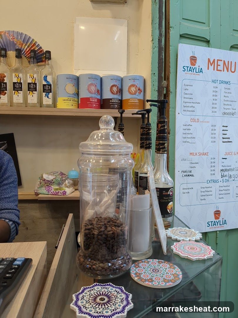 A close-up view of a coffee shop counter featuring a glass jar filled with roasted coffee beans, decorative coasters, and bottles of syrup or flavoring. In the background, shelves display packaged coffee products and liquor bottles labeled '183'. A menu board on the right lists hot drinks like espresso (₹20), latte (₹35), and milkshakes such as vanilla (₹40) and chocolate (₹40). The setting suggests an inviting café environment with warm lighting and organized product displays.