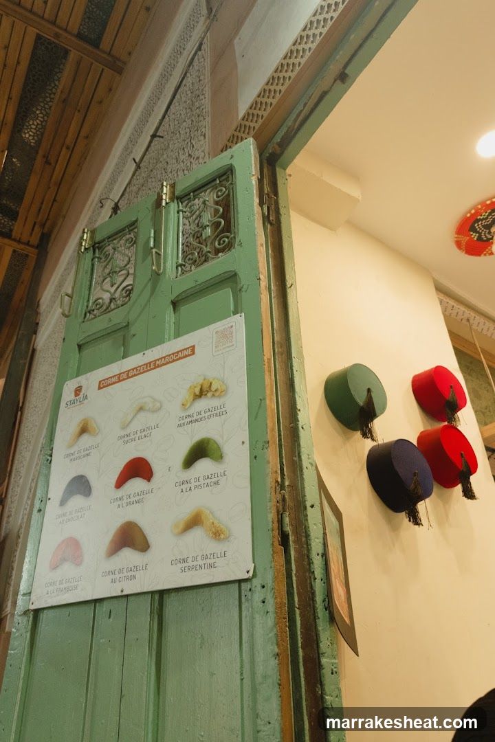 A photo showing an interior space with a green wooden door featuring a menu board titled 'CORNE DE GAZELLE MAROQUINE'. The wall to the right displays colorful hats hanging decoratively.