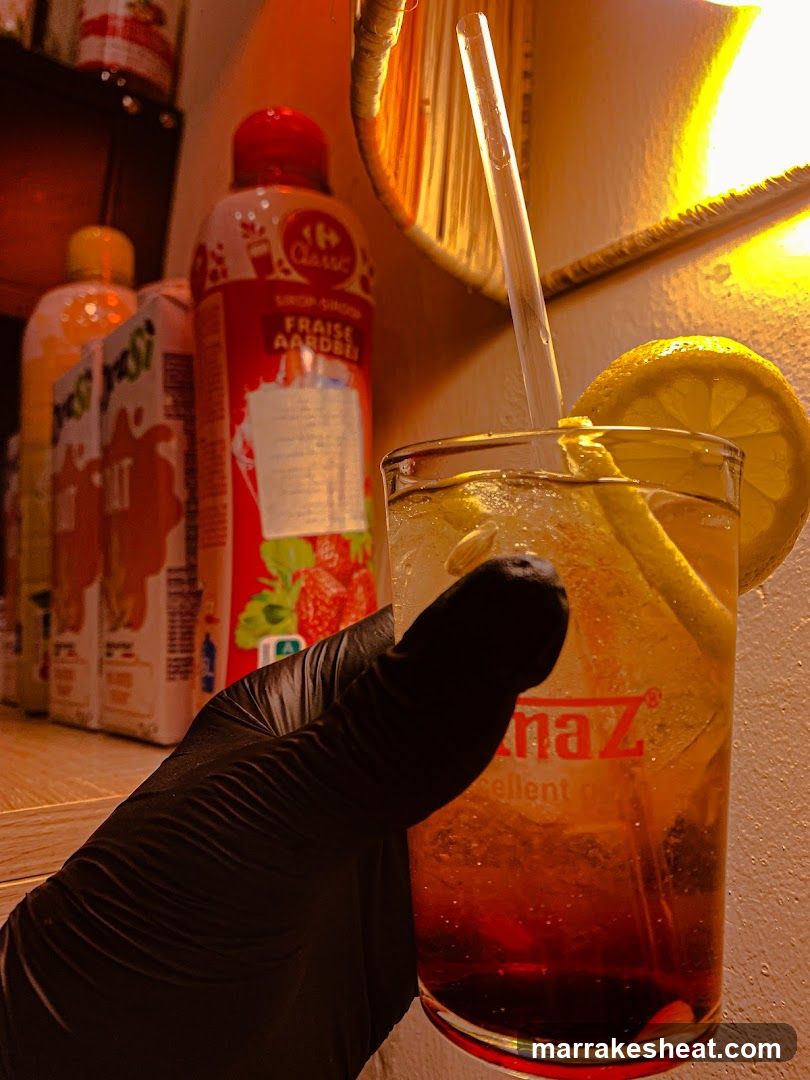 A close-up shot of a hand wearing a black glove holding a glass filled with an amber-colored cocktail, garnished with lemon slices and ice cubes. In the background, there are bottles of fruit juice and other beverages on a shelf, illuminated by warm lighting.