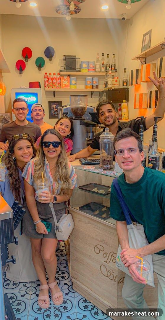 A group of seven people posing inside a cozy coffee shop with colorful decor, including wall-mounted lanterns and shelves stocked with bottles and jars. The counter displays coffee beans in glass containers, and the floor features blue patterned tiles.