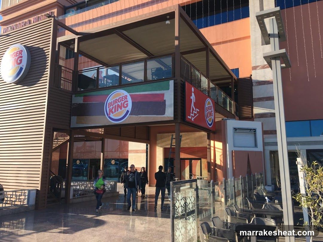 Photo of the physical exterior entrance and storefront facade of a Burger King location. Visible signage includes the Burger King logo on the building wall, a large banner with the Burger King logo and flag design above the entrance, and a red safety sign with a running figure next to another Burger King logo. People are walking near the entrance.