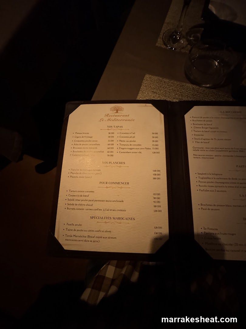 A close-up photo of an open menu from Restaurant La Mediterranee, featuring sections like 'Nos Tapas', 'Nos Planches', 'Pour Commencer', and 'Specialites Marocaines'. The menu is illuminated by ambient lighting in a dimly lit dining setting.